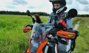 Motocyklista w kasku i kurtce przygotowuje się do podróży motocyklem po Europie. W tle zielone pole i niebo.