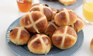 En hög med gyllene, nybakade **hot cross buns** på en blå tallrik, redo att avnjutas med honung och apelsinjuice.