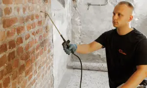 Mann sprüht mit Gerät gegen Feuchtigkeit im Keller. Ziegelwand wird behandelt.