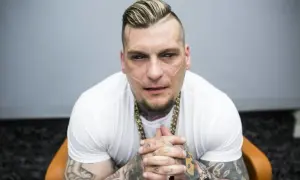 Popek kiedys, z charakterystycznymi bliznami na twarzy i tatuażami, w białej koszulce i złotym łańcuchu, siedzi na krześle.