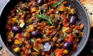 Un plat de caponata sicilienne savoureux, garni d'olives violettes et d'une branche de romarin frais, prêt à être dégusté avec du pain grillé.