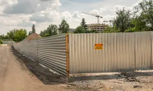 Metalowe ogrodzenie placu budowy z tabliczką "Uwaga, wyjazd z budowy". W tle budynki wznoszone przez dźwigi.