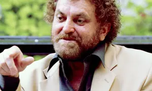 Reżyser z kręconymi włosami i brodą, gestykuluje palcem, ubrany w jasną marynarkę.