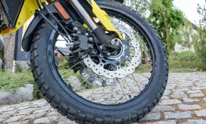 Koło motocykla z tarczą hamulcową i zaciskiem, na tle kamiennej nawierzchni i zieleni.