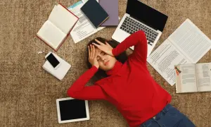 Zmęczona studentka leży na dywanie otoczona książkami, laptopem i telefonem.