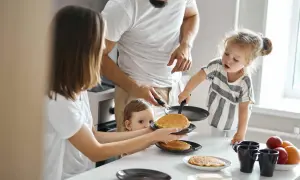 Rodzina przygotowuje placuszki dla niemowlaka 7 miesięcy. Mama podaje talerz z placuszkami, a tata i starsza córka pomagają na patelni.