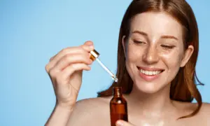 Rudowłosa kobieta z piegami uśmiecha się, trzymając w dłoni buteleczkę z serum i pipetkę. Jej skóra lśni, jakby właśnie nałożyła witaminowy eliksir.