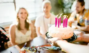 Tort urodzinowy z płonącymi różowymi świeczkami, podawany przez ręce, na tle uśmiechniętych ludzi.