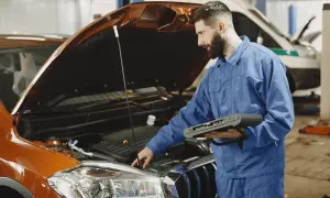 Mécanicien en bleu effectuant un diagnostic électronique sur un moteur de voiture orange. Il tient un appareil de diagnostic, essentiel pour connaître le prix d'un diagnostic voiture.