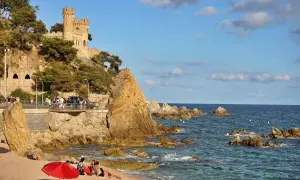 El kasteel Lloret de Mar se alza sobre una colina rocosa junto a una playa con gente disfrutando del sol y el mar azul.