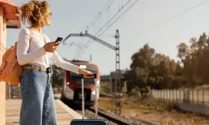 Kobieta z plecakiem i walizką na peronie sprawdza telefon, zastanawiając się, ile się płaci za nadbagaż. Pociąg zbliża się do stacji.