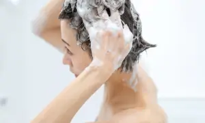 Une femme se lave les cheveux sous la douche, la mousse du shampoing coule sur son visage. Elle masse son cuir chevelu, soulageant la transpiration.