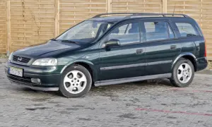 Zielony Opel Astra kombi, jeden z wielu aut w ocynku, stoi na mokrym parkingu przed drewnianym płotem.