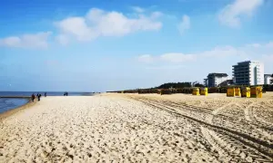 Ein weiter, sonniger Duhnen Strand mit gelben Strandkörben und einigen Spaziergängern. Im Hintergrund moderne Gebäude.