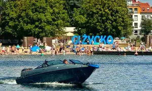 Łódź motorowa płynie po jeziorze, w tle plaża z ludźmi i napis "GIŻYCKO". Idealne miejsce, gdzie na Mazury!