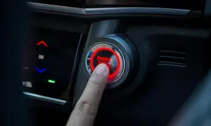 Un doigt appuie sur le bouton "ENGINE START" qui clignote en rouge. La voiture est prête à démarrer.