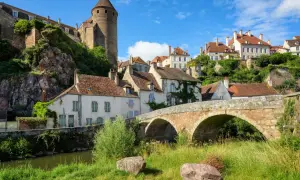 Un village pittoresque de la Bourgogne avec un pont de pierre ancien, des maisons aux toits rouges et une tour médiévale perchée sur une colline verdoyante.