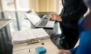Plan mieszkania i laptop na blacie. Dwie osoby omawiają szczegóły, być może dotyczące ubezpieczenia mienia ruchomego.