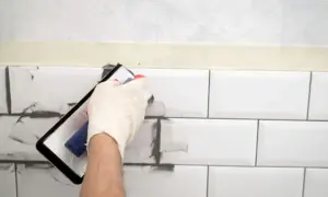 Ręka w rękawiczce nakłada szarą masę na białe płytki, pokazując, czym pomalować fugi.