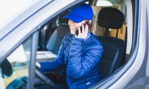Kobieta w niebieskiej czapce i kurtce siedzi za kierownicą i rozmawia przez telefon.