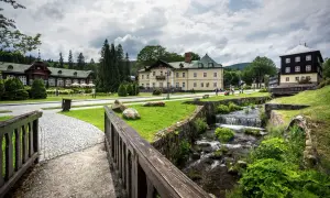 Idyllische Szene in Karlova Studanka: Gebäude im Kurort, ein Bach plätschert über Steine, umgeben von Grün.