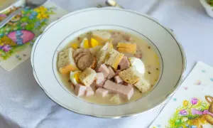 Biała zupa z kiełbasą, jajkiem i chlebem w talerzu, obok serwetki z zajączkiem.