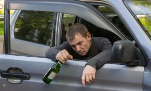 Mężczyzna z butelką w ręku opiera się o otwarte drzwi samochodu. Jego mina sugeruje, że może być to jazda pod wpływem.