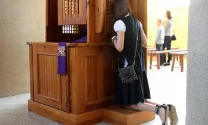 Dziewczyna klęczy przy konfesjonale, kiedy wprowadzono spowiedź na ucho. W tle widać ludzi czekających na ławkach.