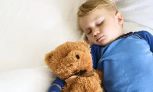 En liten pojke sover sött med sin nallebjörn. Hur många timmar ska en 10-åring sova? Sömn är viktigt för barn.