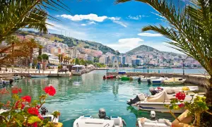 Albania, gdzie najpiękniej! Widok na port z palmami, kolorowymi budynkami na wzgórzu i łodziami na turkusowej wodzie.