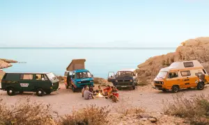 Vier bunte Campervans am Meer, Menschen sitzen am Lagerfeuer.