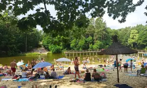 Słoneczny dzień na plaży przy zalewie w Garbatce. Ludzie odpoczywają na piasku, kąpią się w wodzie i spacerują po pomoście.