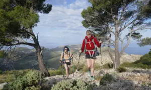 Zwei Wanderinnen mit Rucksäcken und Stöcken auf einem felsigen Pfad, umgeben von Bäumen und mit Blick auf eine hügelige Landschaft und das Meer. Eine tolle Tour, wie auf komoot geplant.