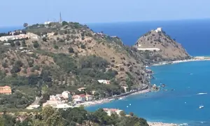 Panoramica di Barcellona Pozzo di Gotto con case bianche e rosse affacciate sul mare blu intenso, colline verdi e un castello in cima.