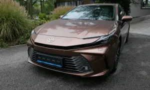 Nowa Toyota Camry 2024 w kolorze brązowym, z kroplami deszczu na karoserii, zaparkowana przed nowoczesnym budynkiem.