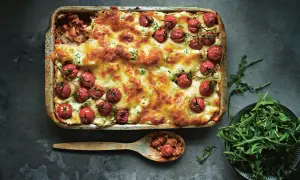 En gyllene salsiccia lasagne toppad med körsbärstomater och ost. En sked med lasagne och en skål rucola bredvid.