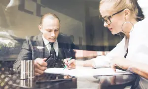 Mężczyzna i kobieta omawiają dokumenty. Kobieta z okularami i kolczykami-kołami pisze, mężczyzna trzyma długopis. Być może podpisują pełnomocnictwo.