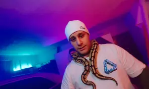 Macias w białej czapce i t-shircie z wężem na szyi, w neonowym świetle.