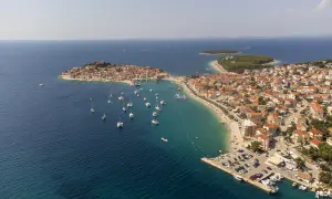 Malownicze miasteczko na wybrzeżu Chorwacji, gdzie turkusowe morze otacza wyspę z czerwonymi dachami. Wiele łodzi cumuje przy brzegu, a plaża tętni życiem.