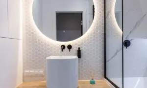 Moderno baño con lavabo blanco, espejo redondo iluminado y azulejos hexagonales. Ideal para tu presupuesto baño.