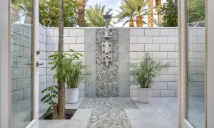 Doccia esterna in muratura con finiture in ciottoli e piastrelle bianche. Piante verdi e palme sullo sfondo creano un'oasi tropicale.