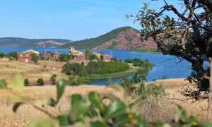 Vue sur le lac du Salagou, avec ses eaux bleues, ses collines rouges et les ruines d'un village. Un voilier navigue paisiblement.