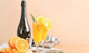 Dwa kieliszki orzeźwiającego mimoza drinka z plasterkami pomarańczy i gałązką rozmarynu, obok butelka szampana i shaker.