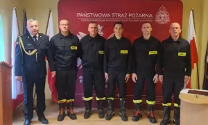 Sześciu strażaków w mundurach, w tym dowódca, tworzy rota strażacka. Stoją na tle sztandarów i logo Państwowej Straży Pożarnej.