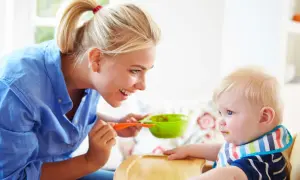 Mama karmi dziecko łyżeczką z zielonej miseczki. Dziecko ma śliniaczek, a mama uśmiecha się do niego. To posiłek bogaty w błonnik dla dziecka.