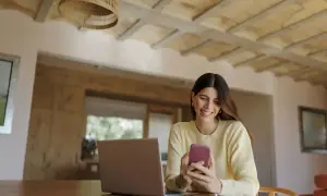 Uśmiechnięta kobieta korzysta z telefonu, planując przelewy w mBanku. Obok laptop i notatnik.
