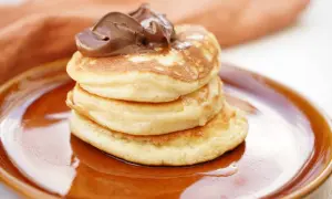 En hög med fluffiga amerikanska pannkakor toppas med chokladkräm på en brun tallrik.