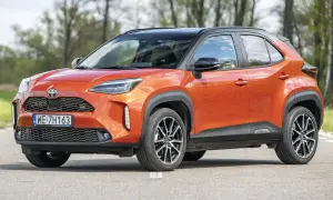 Pomarańczowy Toyota Yaris Cross 2024 z czarnym dachem, zaparkowany na drodze, z drzewami w tle.