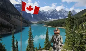 Persona con mochila admira el lago turquesa y las montañas nevadas de Canadá. La bandera canadiense ondea, invitando a una aventura de work and holiday Canada.