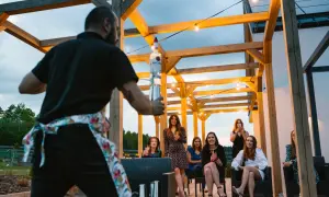 Barman przygotowuje drinki dla grupy kobiet, świetne tanie pomysły na wieczór panieński pod drewnianą pergolą z lampkami.